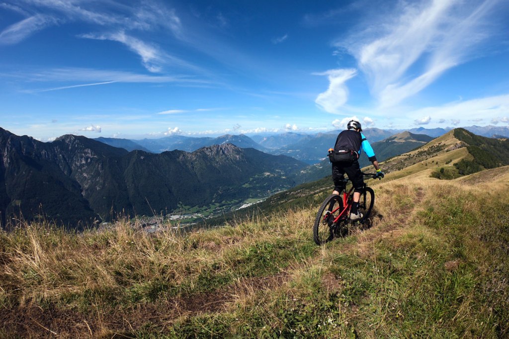 un ciclista scende con la mountain bike lungo una cresta erbosa con panorama sulle montagne lombarde e le valli