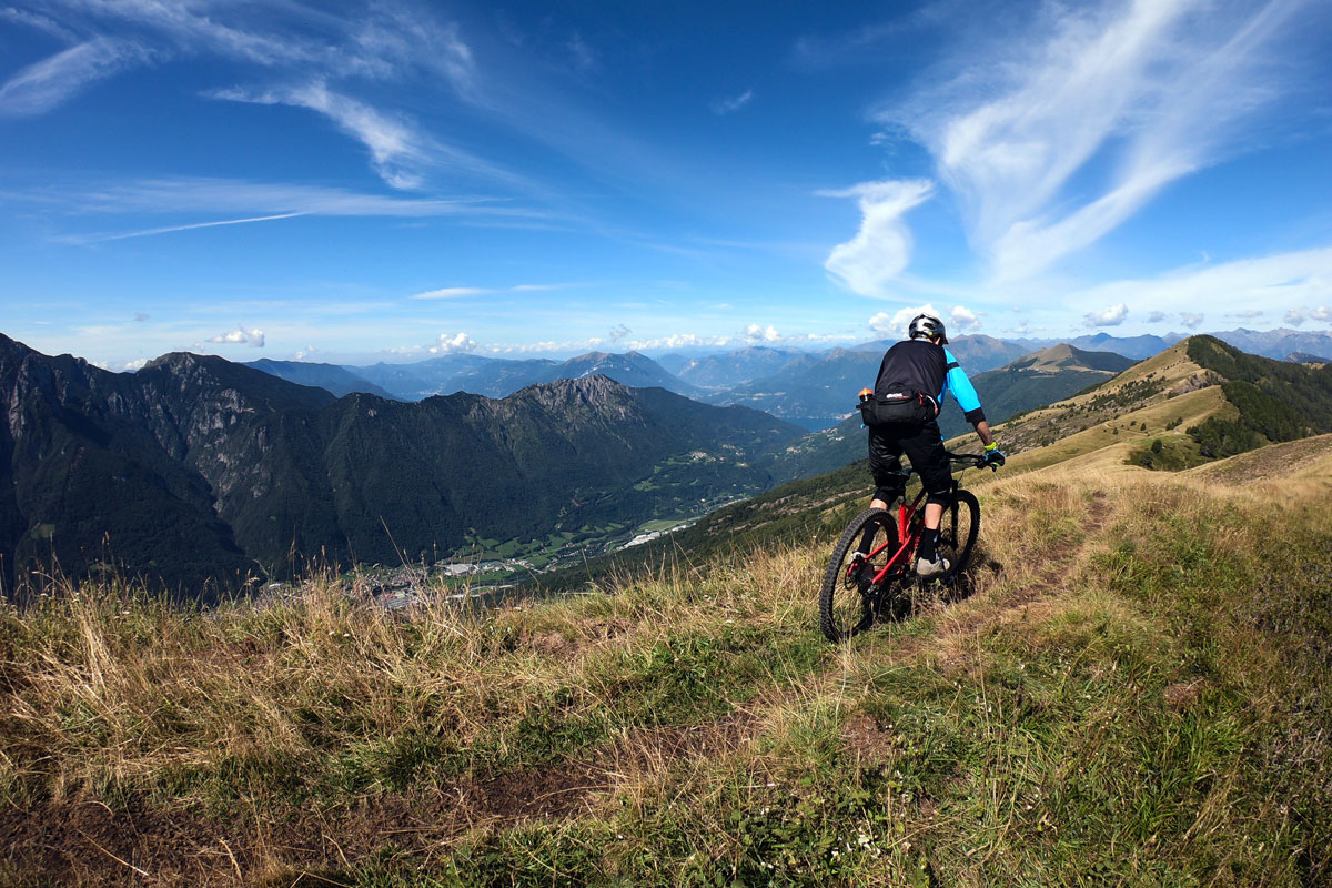un ciclista scende con la mountain bike lungo una cresta erbosa con panorama sulle montagne lombarde e le valli