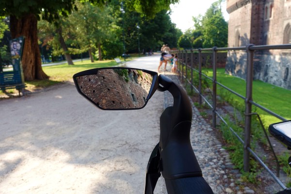 specchietto retrovisore in primo piano alberi e una strada di ghiaia in secondo piano