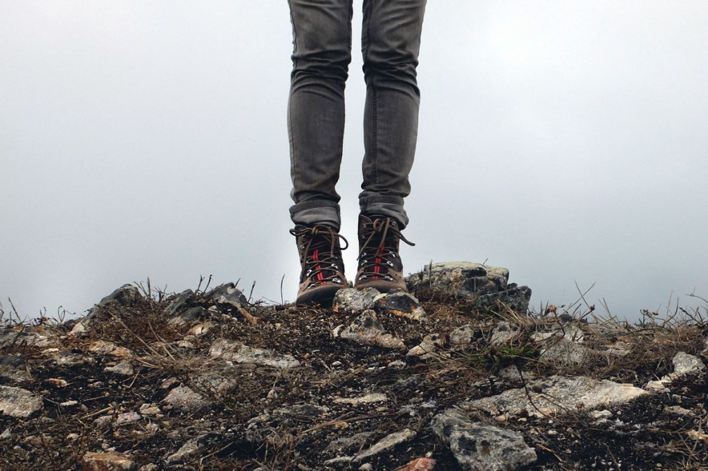 terreno brullo di montagna in primo piano e in secondo piano delle gambe e degli scarponi da montagna con nebbia sullo sfondo