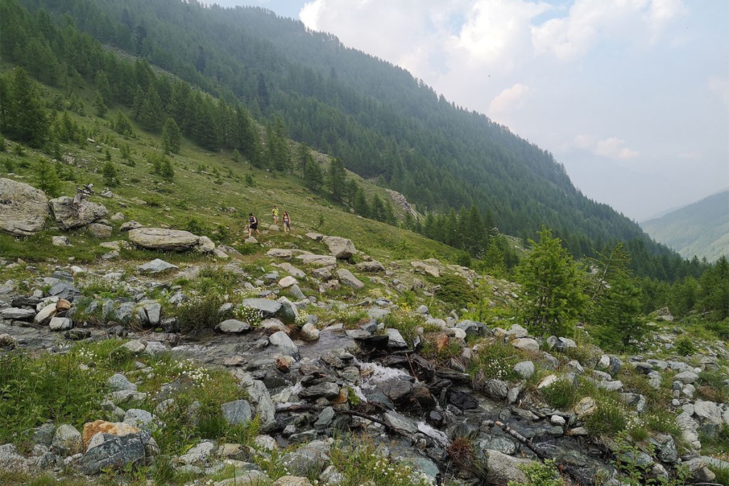 un paesaggio di montagna con un torrente in primo piano, alcuni escursionisti a piedi e la valle sullo sfondo