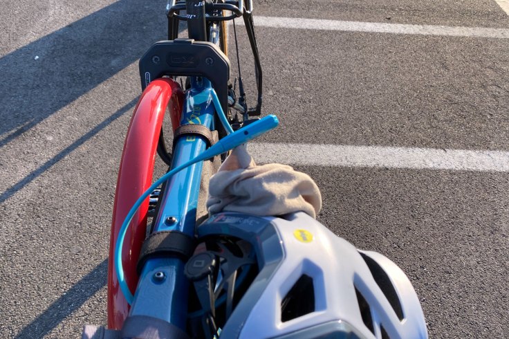 in primo piano un lucchetto a fascetta assicura il casco ed una bandana in lana merino alla bicicletta. Sullo sfondo un lucchetto del tipo U Lock assicura la bicicletta ad un paletto in ferro