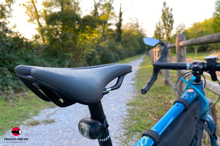sella per bicicletta grigio scuro in primo piano ed un sentiero di ghiaia in secondo piano. la bicicletta nella foto è equipaggiata anche con specchietto retrovisore
