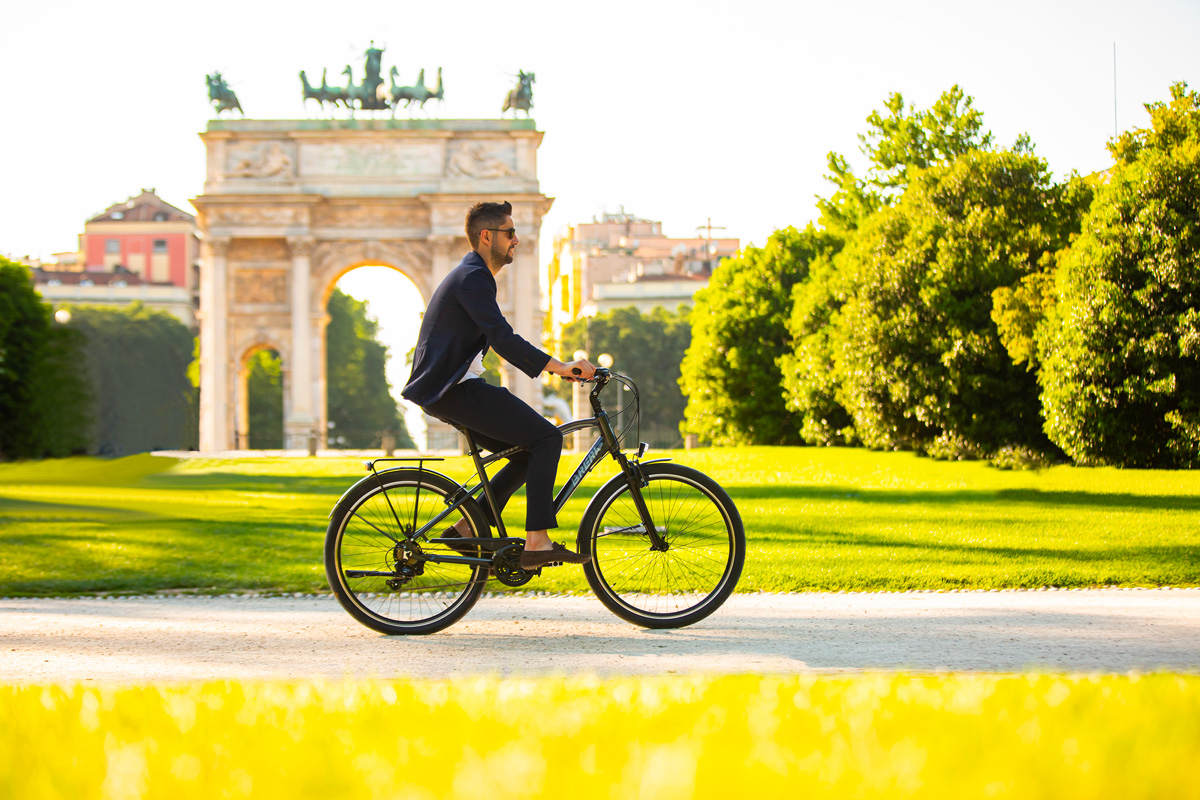 un ragazzo in giacca e tenuta da ufficio attraversa il Parco Sempione a Milano in sella alla sua bicicletta nei pressi dell'Arco della Pace