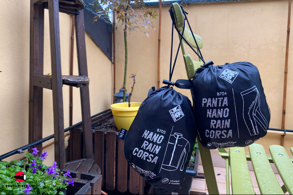 in primo piano un completo antipioggia per ciclismo racchiuso all'interno della pratica tasca che lo contiene quando non in utilizzo e sullo sfondo la talea di una rosa un acero giapponese e altre piante su un balcone milanese