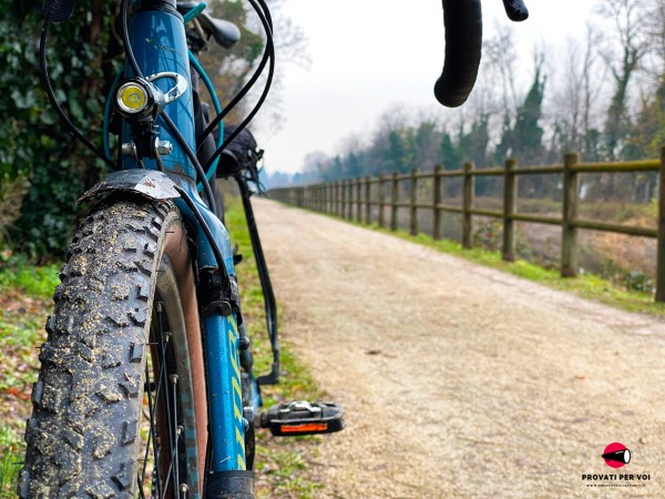 primo piano sullo pneumatico gravel bike montato su una bici poggiata ad un albero sullo sfondo una pista ciclabile in ghiaia che costeggia il Canale Villoresi
