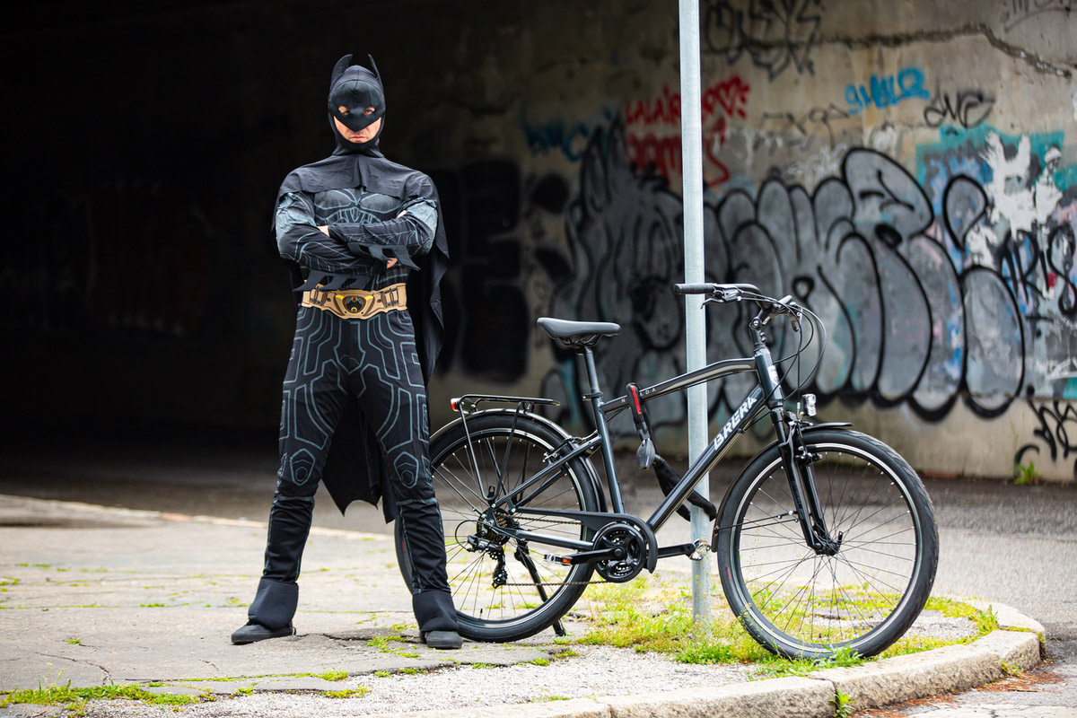 un uomo mascherato da Batman posa vicino ad una bicicletta legata ad un paletto con un lucchetto a catena nei pressi di un sottopasso ferroviario della periferia milanese