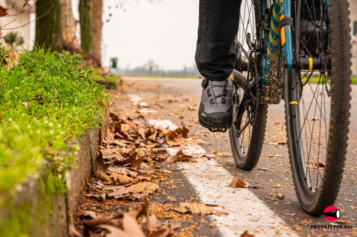 un paio di scarpe per il ciclismo invernale fotografate in ambiente tra foglie secche autunnali, erbetta verde che sa di primavera e una biciclette
