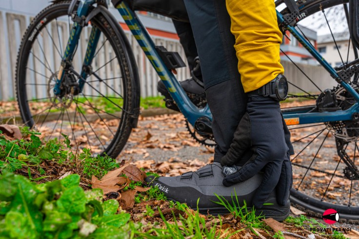 un paio di scarpe per il ciclismo invernale fotografate in ambiente tra foglie secche autunnali, erbetta verde che sa di primavera e una biciclette