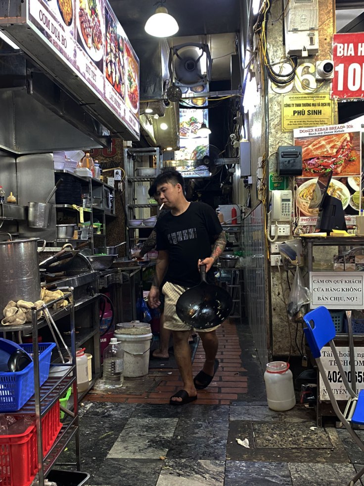 un uomo in pantaloni corti e ciabatte da piscina con una pentola Wok in mano si accinge a mettere sui fornelli un altro piatto tipico della rinomata cucina vietnamita