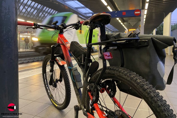 una mountain bike equipaggiata con un portapacchi compatibile con qualsiasi bicicletta e un treno regionale in arrivo al binario