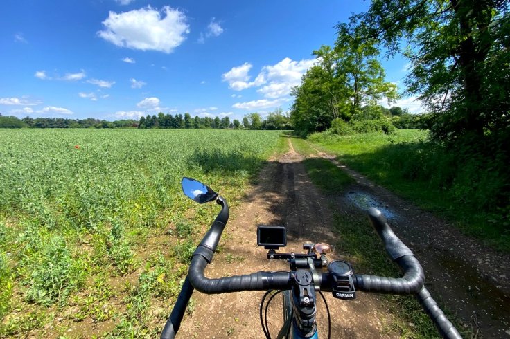 Vista dal manubrio di una bicicletta gravel lungo una strada sterrata tra campi e alberi in Lombardia.